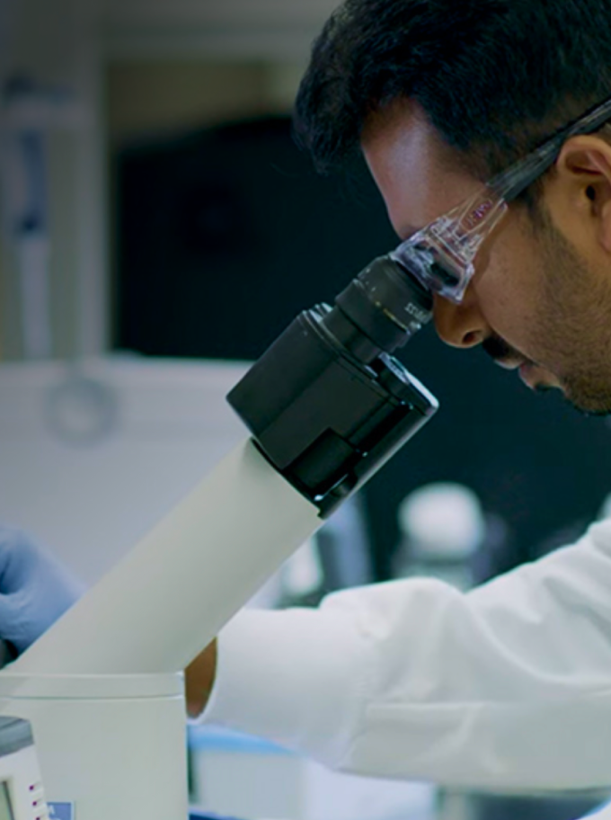 Scientist wearing safety glasses uses a microscope in a laboratory setting for research.